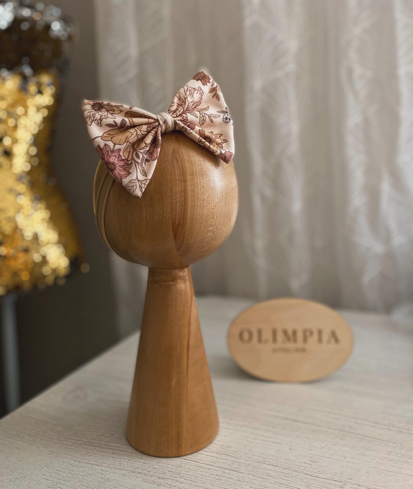 Kwiatowa opaska z kokardą na drewnianej głowie manekina, na tle błyszczącej sukienki i zasłony