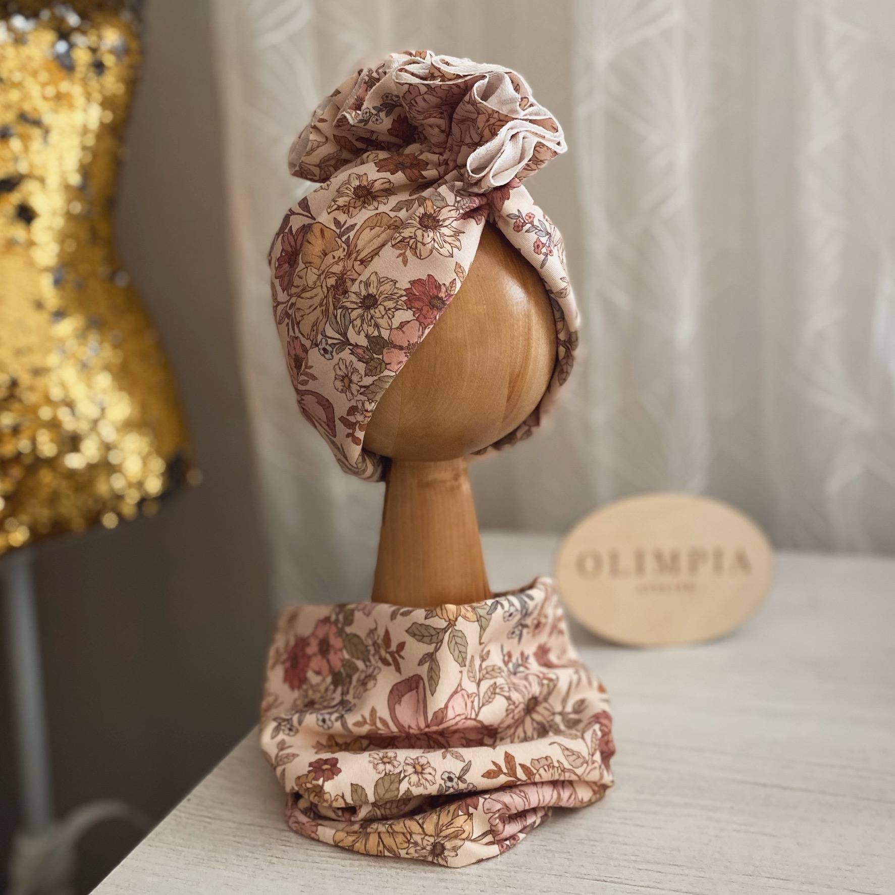 Dziecięca opaska turban z kwiatowym wzorem, prezentowana na drewnianej główce ekspozycyjnej, z dopasowanym szalem na szyję, na tle jasnego wnętrza z manekinem w cekinowej sukience.