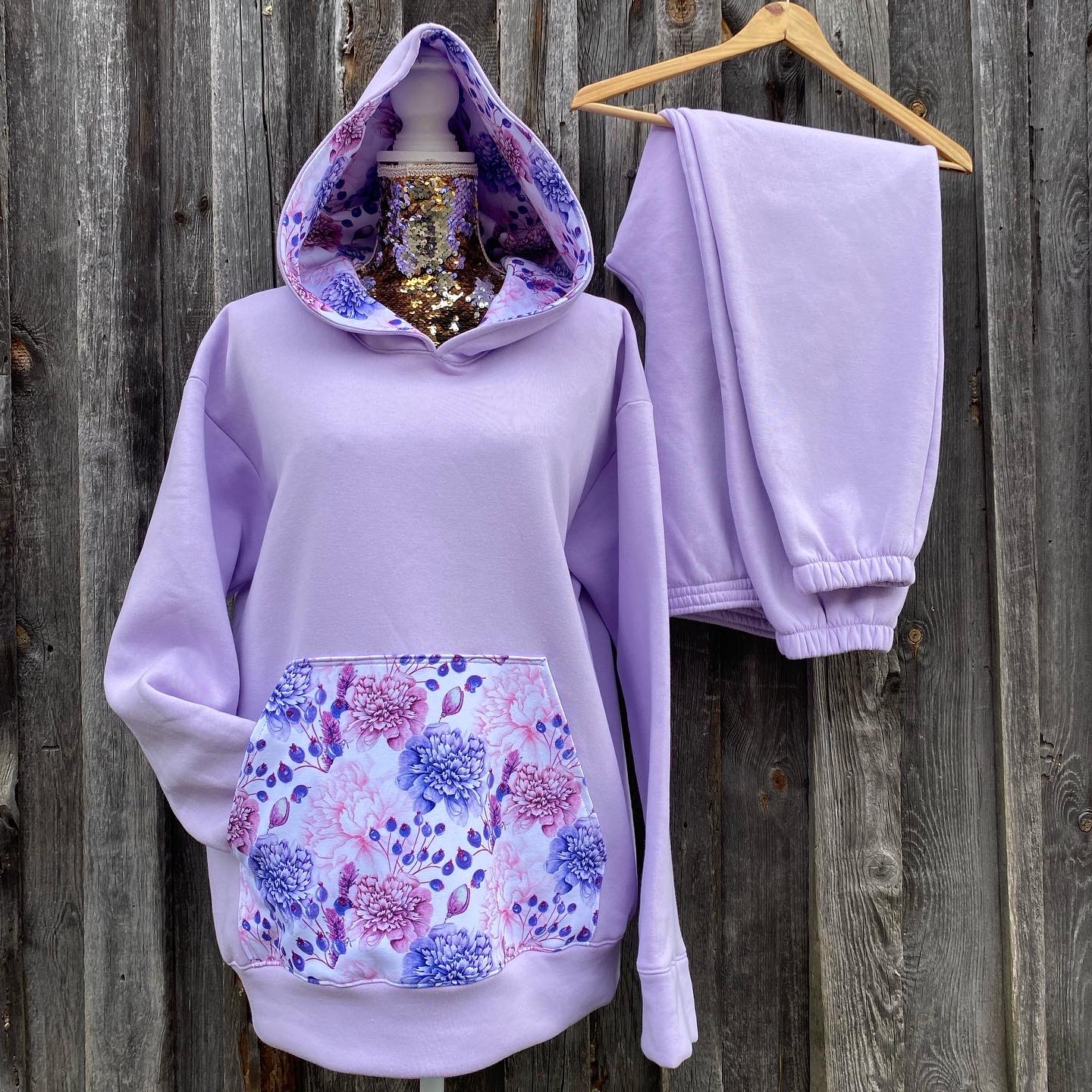 Liliowy komplet dresowy: bluza z kapturem i spodnie, bluza z kieszenią z motywem kwiatowym, całość prezentowana na manekinie i wieszaku na tle drewnianej ściany.