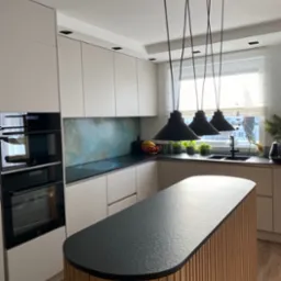 Nowoczesna kuchnia z wyspą o zaokrąglonym blacie z ciemnego kamienia i drewnianą podstawą z pionowych listew, oświetlona trzema czarnymi lampami wiszącymi.