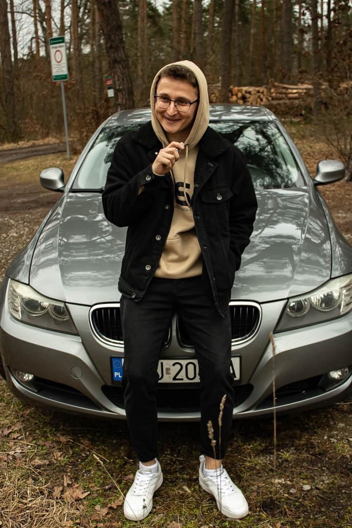 Młody mężczyzna w okularach i bluzie z kapturem oparty o maskę srebrnego samochodu na tle leśnym.