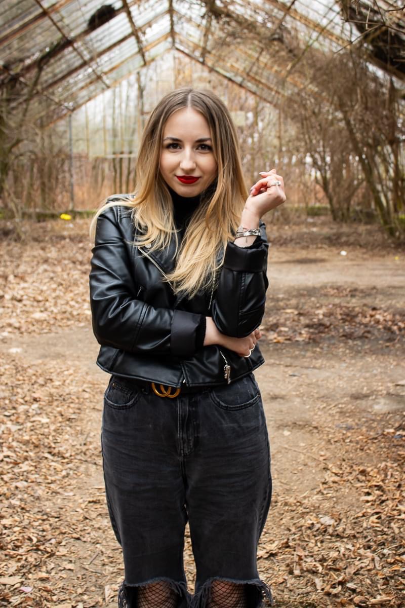Kobieta w czarnej skórzanej kurtce i dżinsach pozuje na tle opuszczonej szklarni, z wyraźnym makijażem ust w kolorze czerwieni i blond włosami.