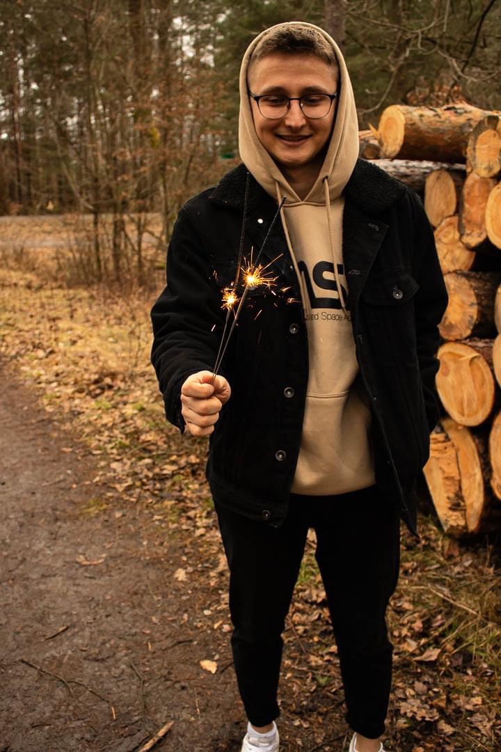 Młody mężczyzna w bluzie z kapturem i kurtce, trzymający w dłoniach płonące zimne ognie na tle leśnej ścieżki i stosu drewna.