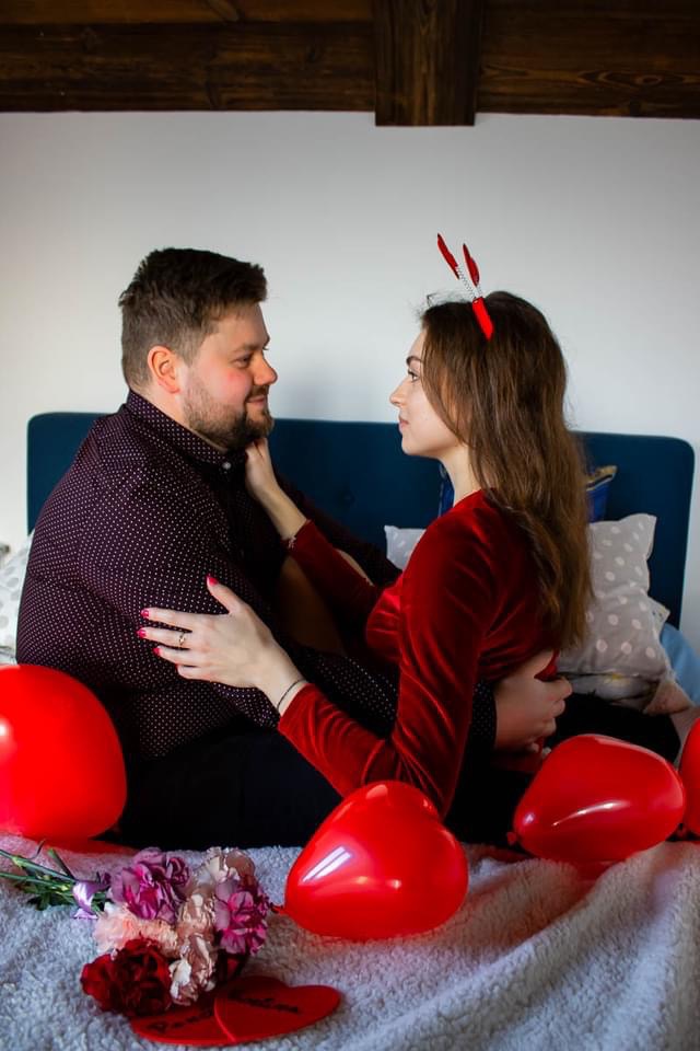 Zakochana para w Opolu Lubelskim podczas sesji narzeczeńskiej, ona w czerwonej sukience z opaską z rogami diabła, on w ciemnej koszuli w kropki, siedzą na łóżku z czerwonymi balonami i kwiatami.