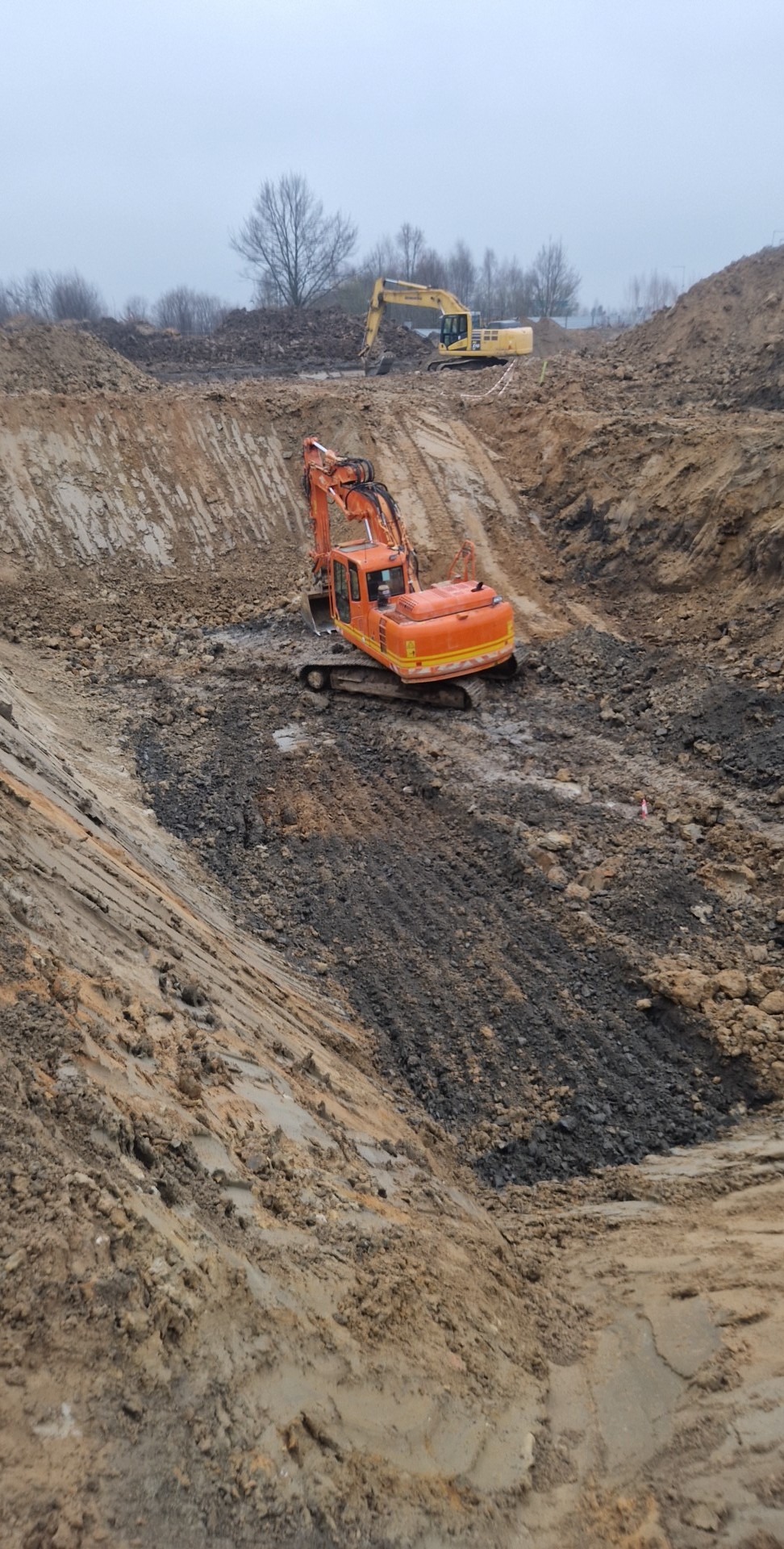 Pomarańczowa koparka gąsienicowa w wykopie, na drugim planie żółta koparka, pochmurne niebo.