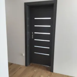 Ciemnoszare drzwi wewnętrzne z poziomymi, prostokątnymi przeszkleniami, zamontowane w jasnym pomieszczeniu, widoczny fragment drewnopodobnej podłogi i gniazdko elektryczne na ścianie.