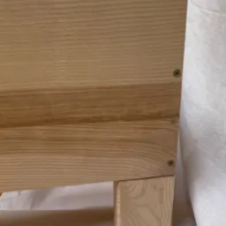 Fragment szafy z jasnego drewna z widocznymi wkrętami i nogą na tle białej tkaniny.