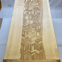 Adam Sułkowski - Blat stołu z jasnego drewna z centralnym pasem dekoracyjnym ułożonym z drobnych, kontrastujących elementów w geometryczny wzór jodełki.