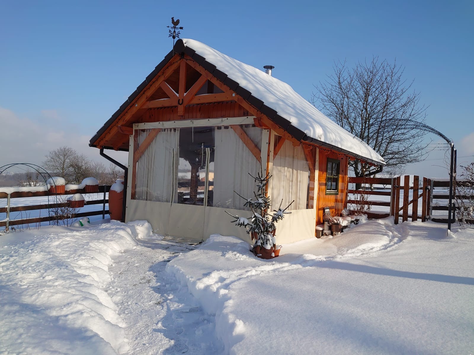 Drewniana altana z zasłonami, pokryta śniegiem, z wiatrowskazem na dachu, otoczona zimowym krajobrazem.