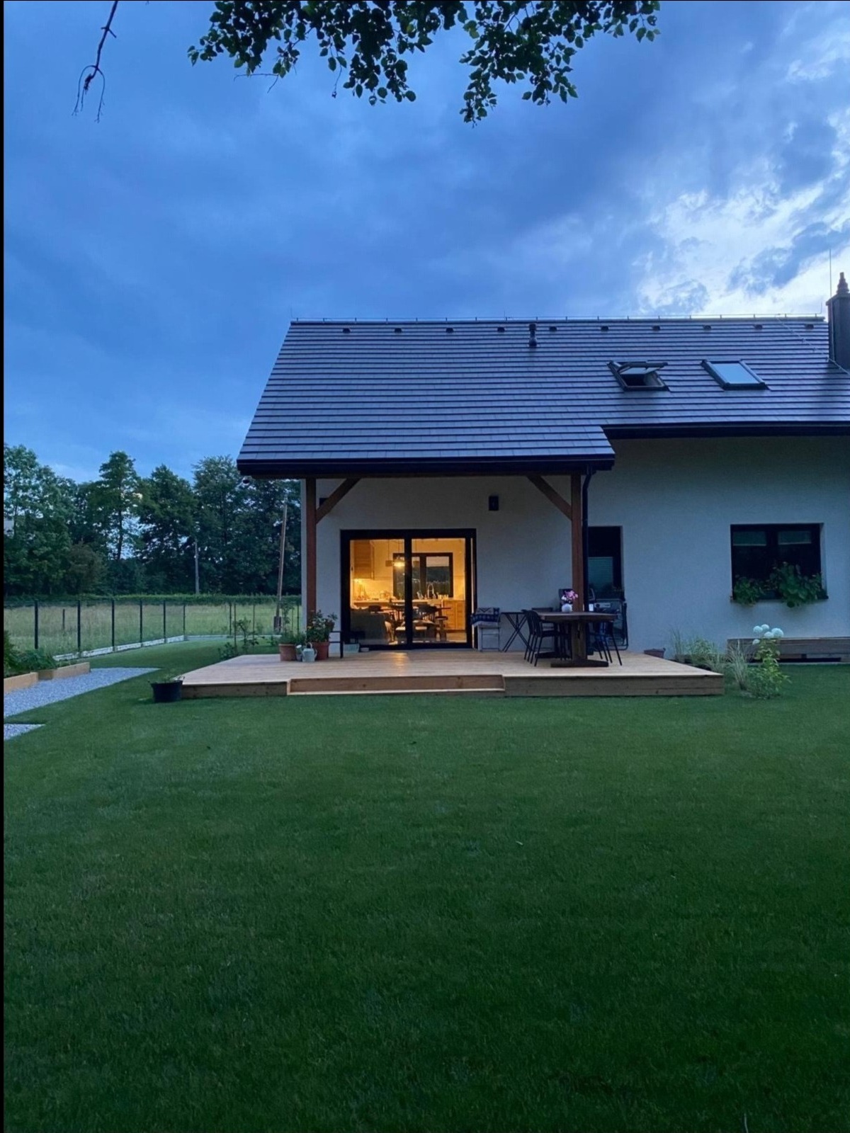 Nowoczesny dom jednorodzinny z drewnianym tarasem i zadaszeniem, widok na ogród w pochmurny dzień, oświetlone wnętrze widoczne przez szklane drzwi.