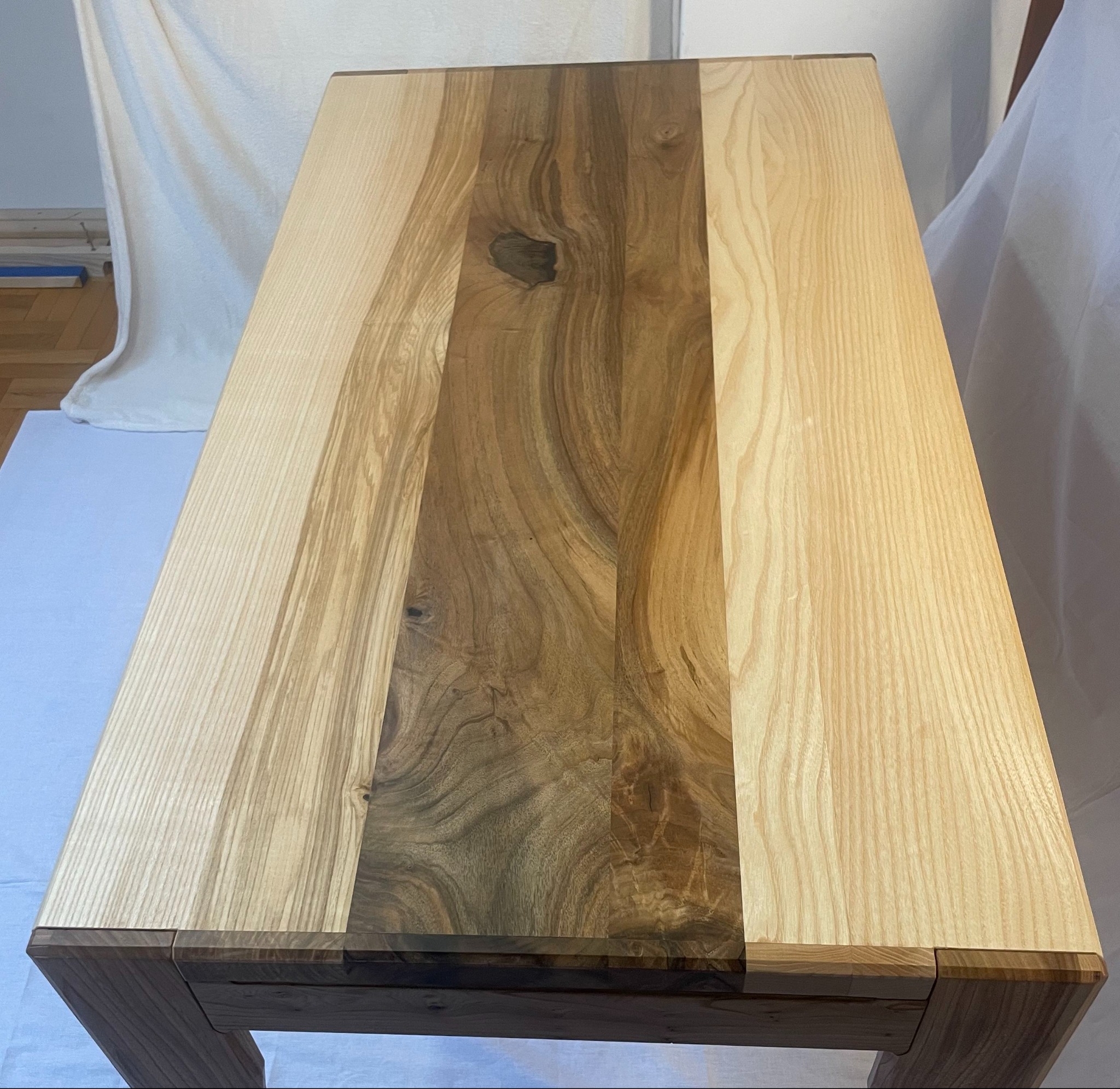 Blat stołu z drewna o różnej barwie i usłojeniu, wykonany w Skoczowie. Widoczne połączenia elementów blatu i fragment nóg stołu. W tle biała tkanina.