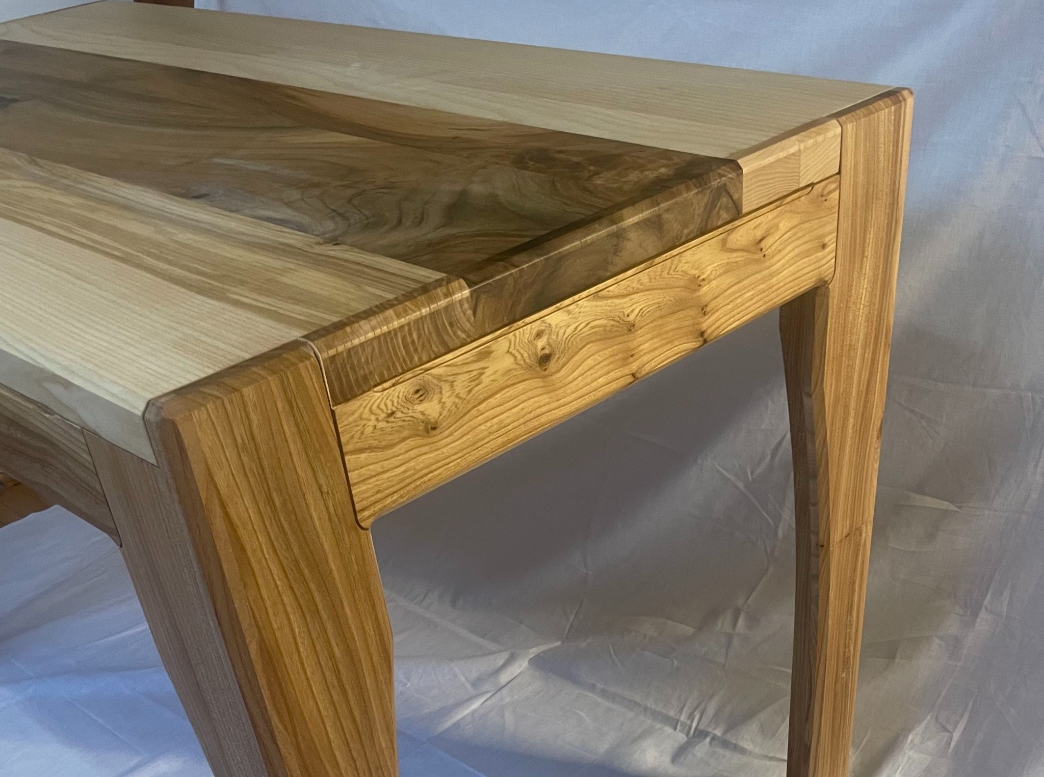 Drewniany stolik z blatem z dwóch rodzajów drewna, z wyraźnym usłojeniem, na toczonych nogach, na tle białej tkaniny.