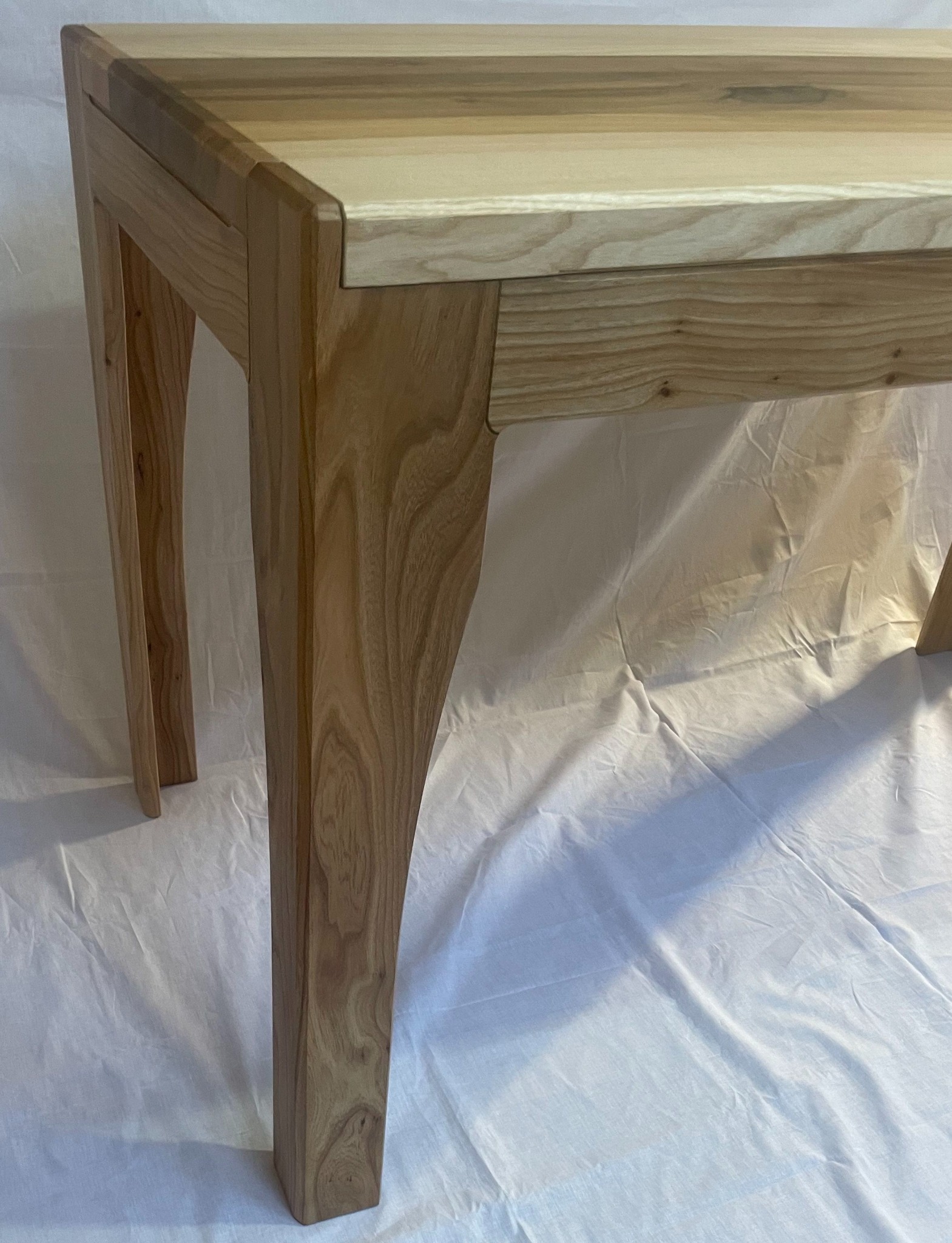 Blat i nogi wykonane z jasnego drewna, z widocznym usłojeniem, na tle białego materiału.