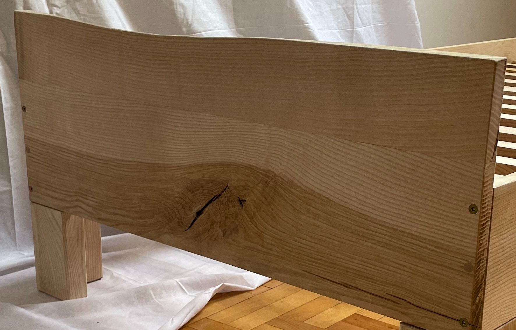 Drewniane wezgłowie łóżka z wyraźnym usłojeniem i sękiem, widoczne śruby mocujące, na tle białej tkaniny i parkietu.