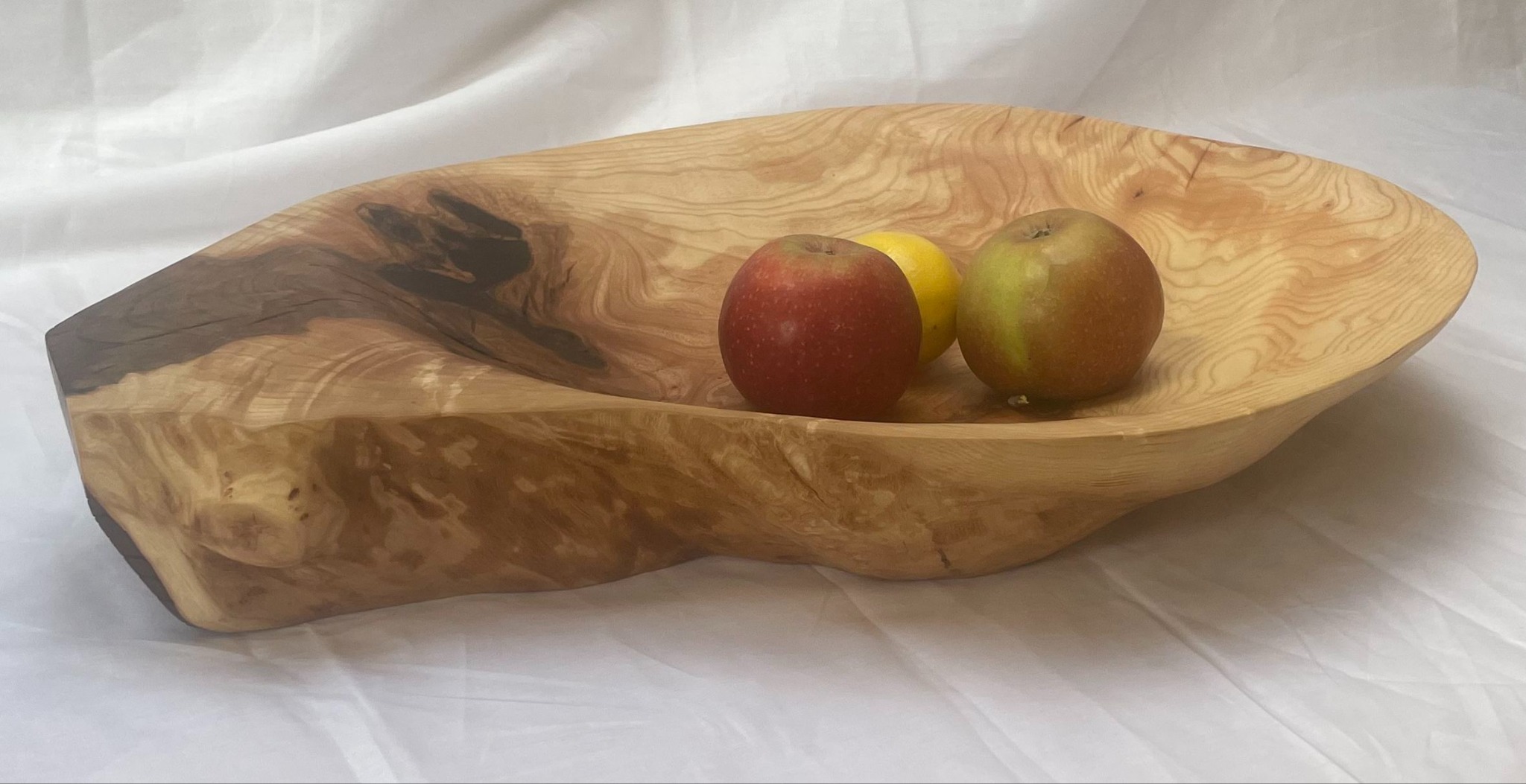 Ręcznie wykonana, nieregularna misa z jasnego drewna z widocznymi słojami, zawierająca jabłko i cytrynę, ustawiona na białym tle.