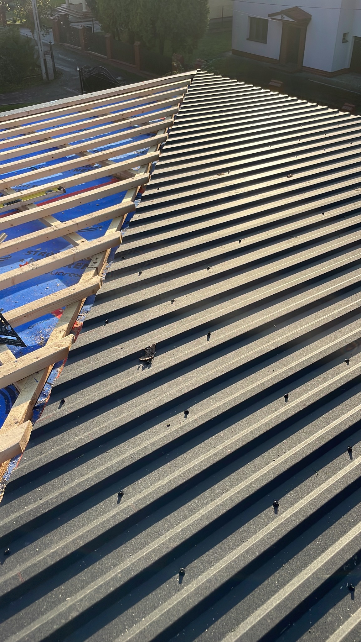 Montaż blachy trapezowej na dachu domu, widoczne drewniane łaty i membrana dachowa, częściowo ukończone pokrycie, widok z góry.