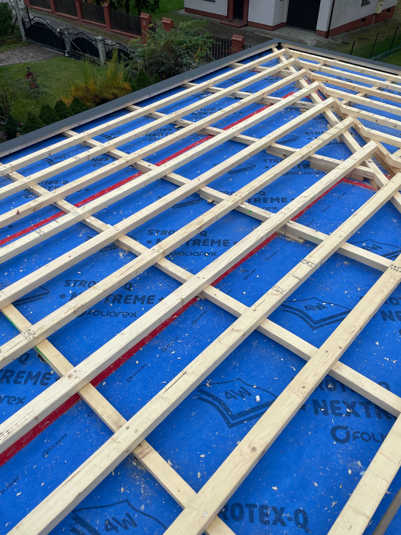 Konstrukcja drewniana dachu w trakcie budowy, widoczne krokwie i łaty na niebieskiej membranie dachowej z nadrukiem, fragment budynku w tle.