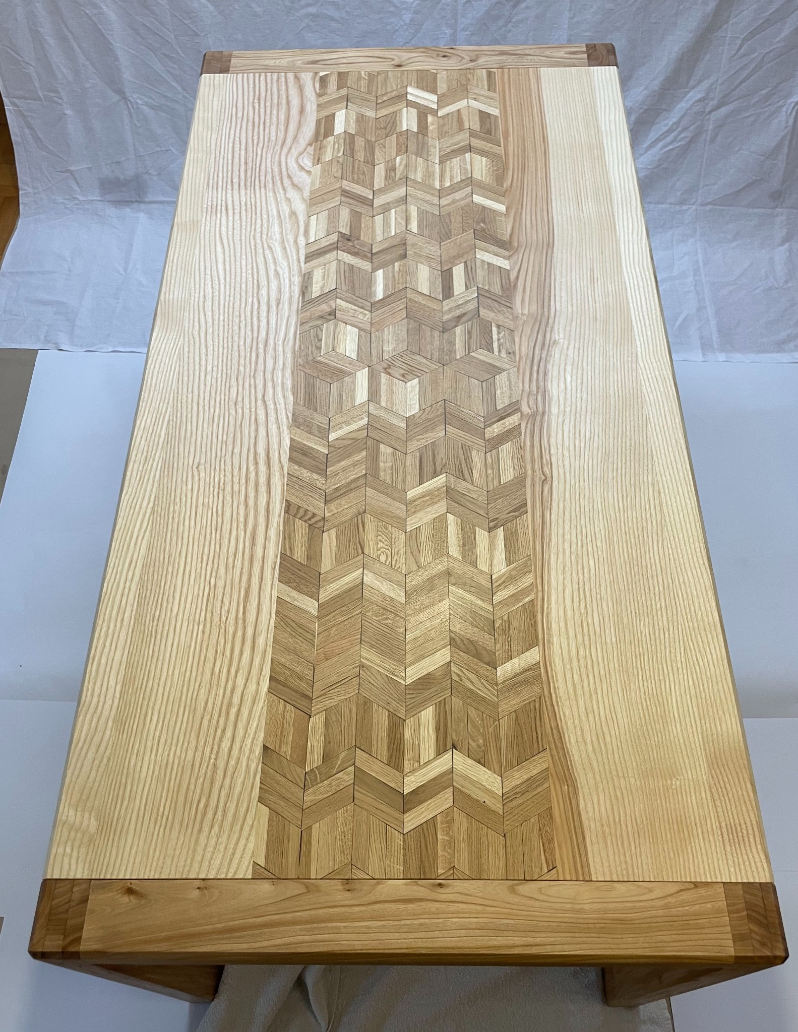 Blat stołu z jasnego drewna z centralnym pasem dekoracyjnym ułożonym z drobnych, kontrastujących elementów w geometryczny wzór jodełki.