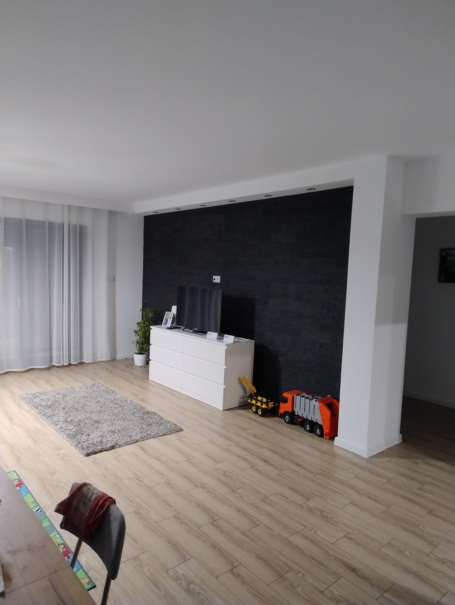 Salon po remoncie z czarną ścianą z imitacji kamienia, jasnymi panelami podłogowymi, białą komodą, szarym dywanem i zabawkami dziecięcymi.