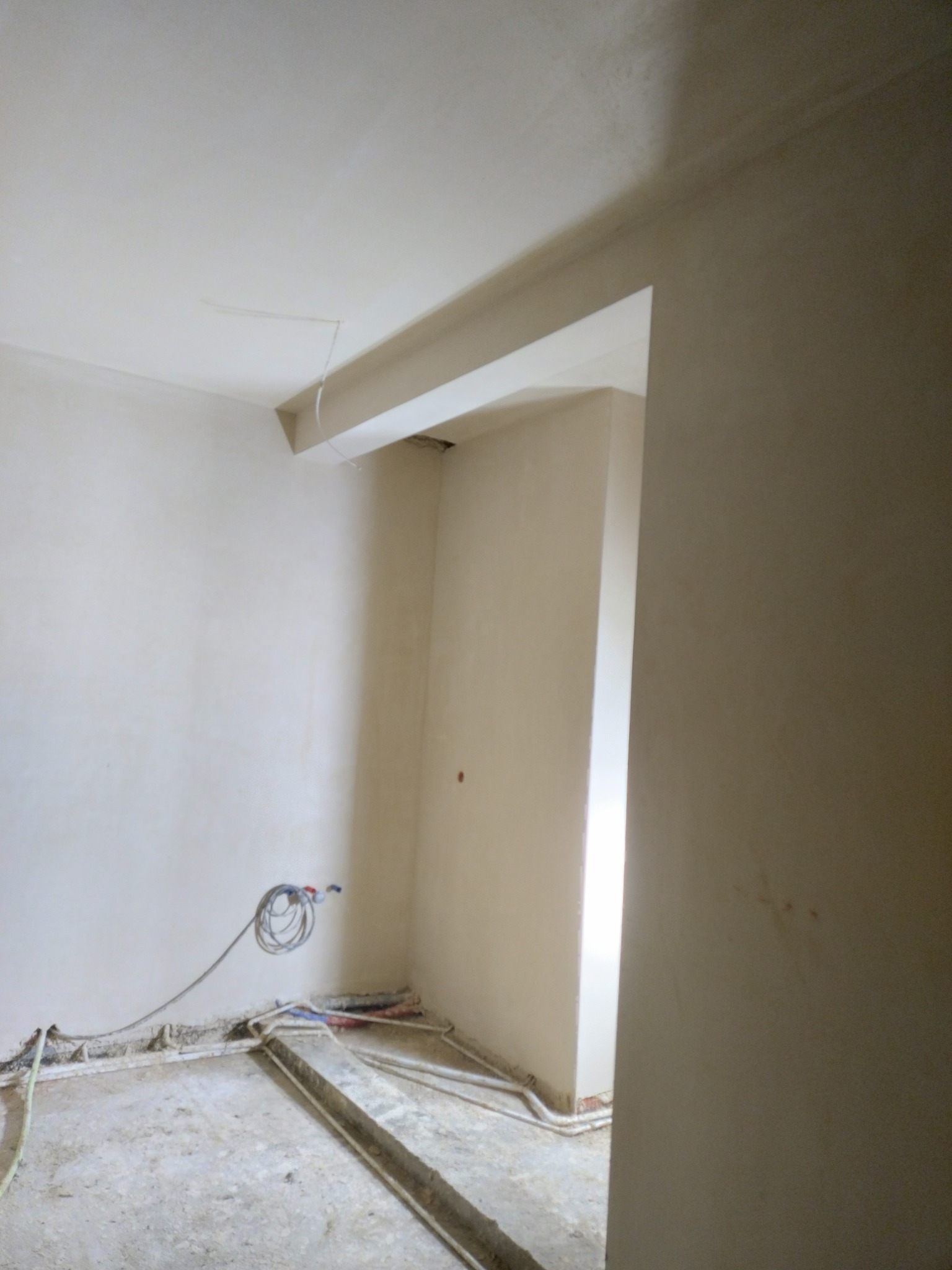 Pokój w trakcie prac wykończeniowych: gładkie, jasne ściany, widoczne przewody elektryczne i rury w podłodze, surowy beton na podłodze.