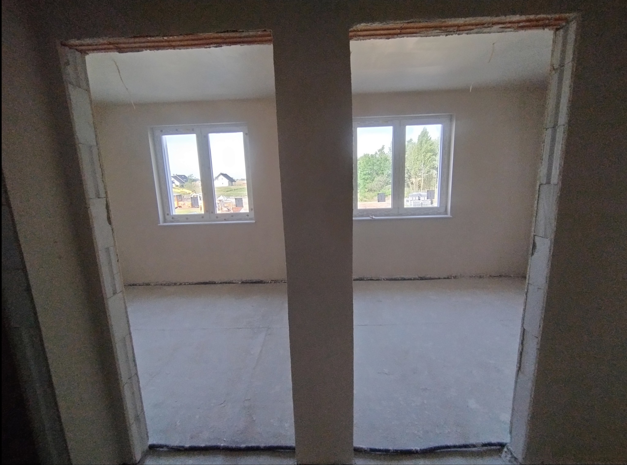 Surowy stan wnętrza z dwoma oknami, widok przez surowy otwór drzwiowy z wyraźnie widocznymi śladami po szpachlowaniu na ścianach i podłodze.