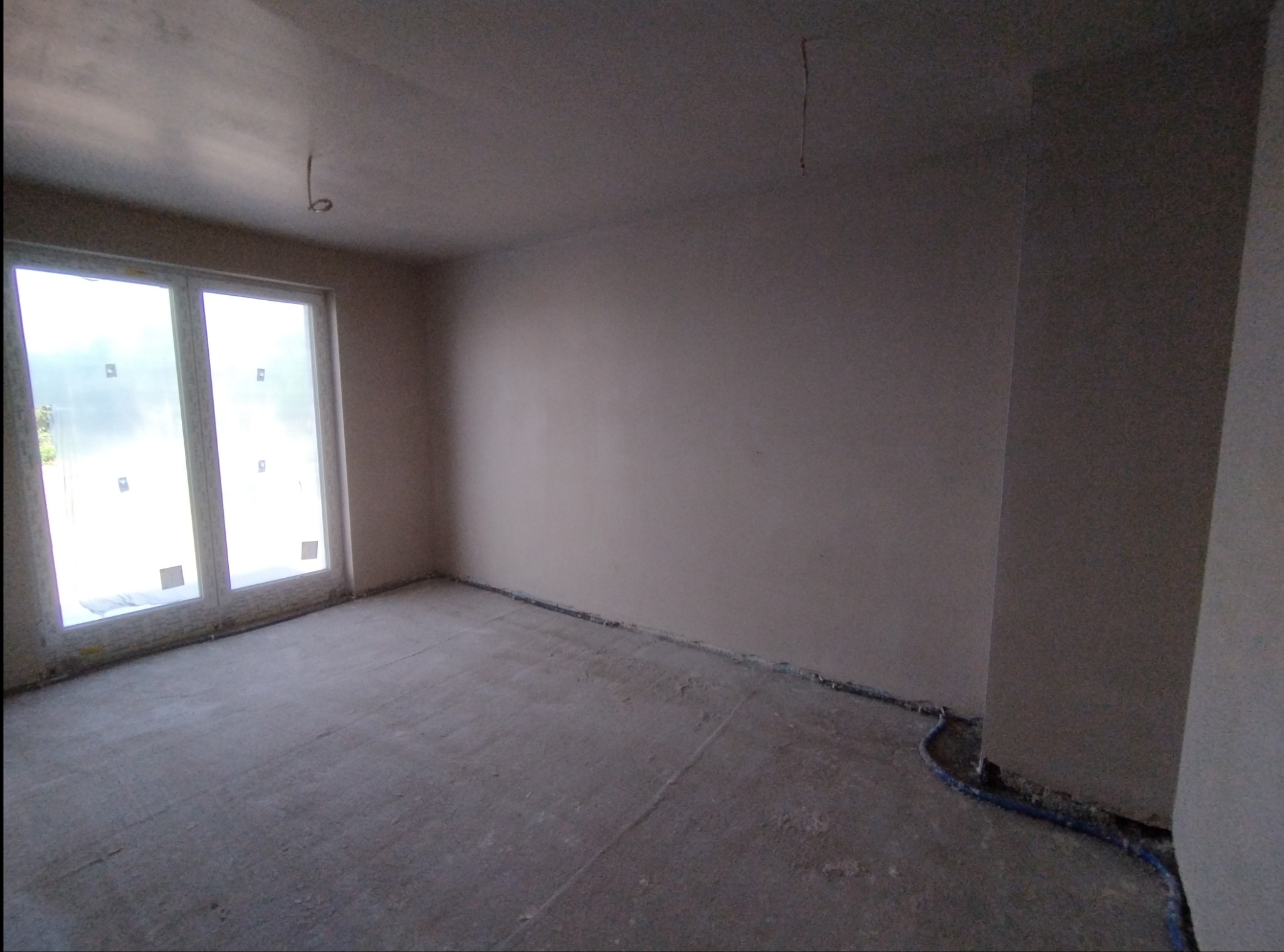 Pokój po nałożeniu gładzi, z widocznymi przewodami elektrycznymi przy podłodze i oknem zabezpieczonym folią.