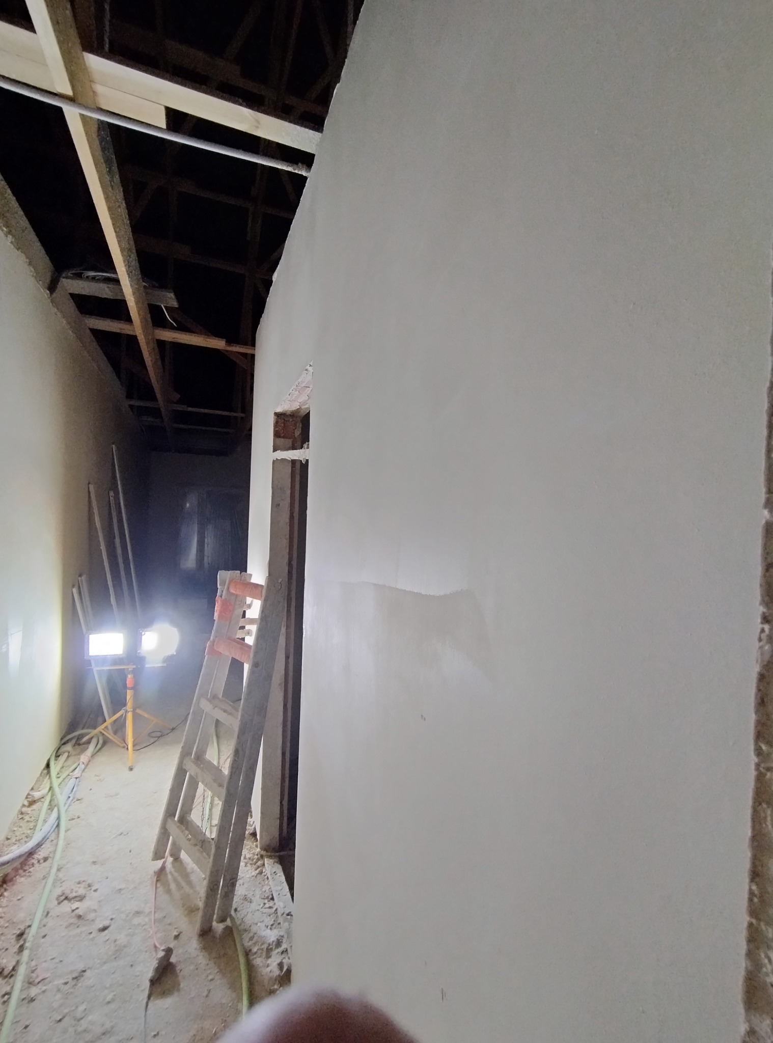 Świeżo położone gładzie na ścianie w trakcie remontu, widoczna drabina, lampa oświetlająca oraz surowy strop budynku.