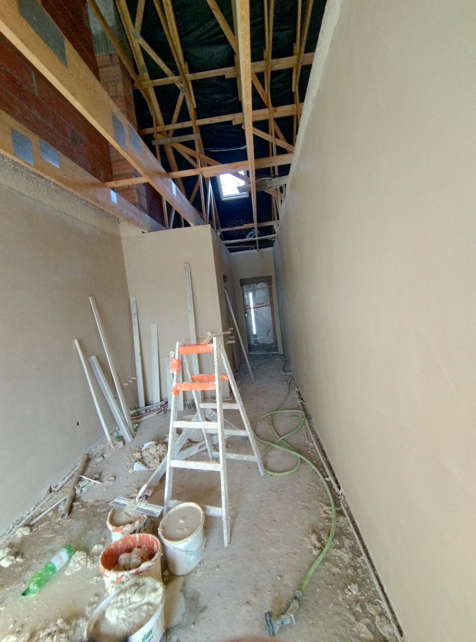 Wnętrze w trakcie prac wykończeniowych: ściany pokryte świeżo nałożoną gładzią, widoczne drewniane elementy konstrukcji dachu, drabina malarska i wiadra z zaprawą na podłodze.