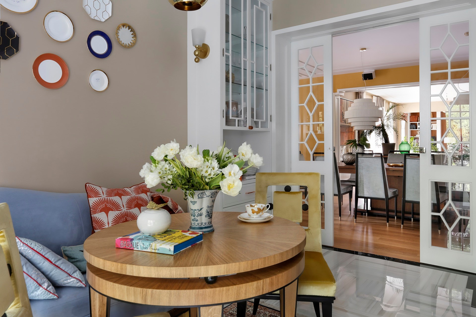 Stylowo urządzona jadalnia z okrągłym drewnianym stołem, bukietem białych kwiatów w wazonie, filiżanką na talerzyku, książką i dekoracyjnymi talerzami na ścianie, widokiem na salon przez...