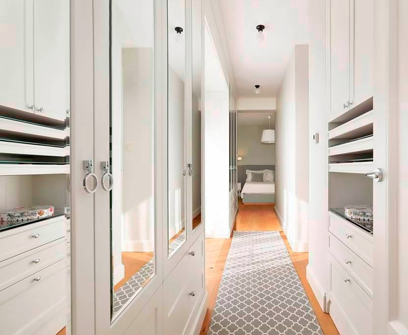 Wnętrze garderoby z białymi szafami wnękowymi z lustrzanymi frontami, widok na korytarz z drewnianą podłogą i dywanem w geometryczny wzór prowadzący do sypialni.