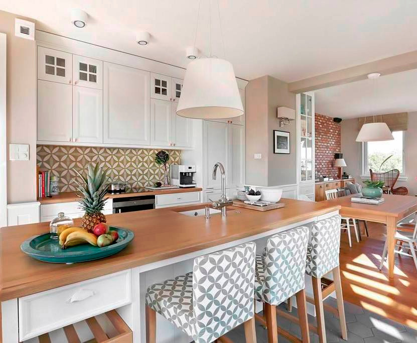 Jasna kuchnia z drewnianym blatem wyspowym, białymi szafkami z małymi okienkami u góry i wzorzystą mozaiką nad blatem, oświetlona wiszącymi lampami i z owocami na blacie.