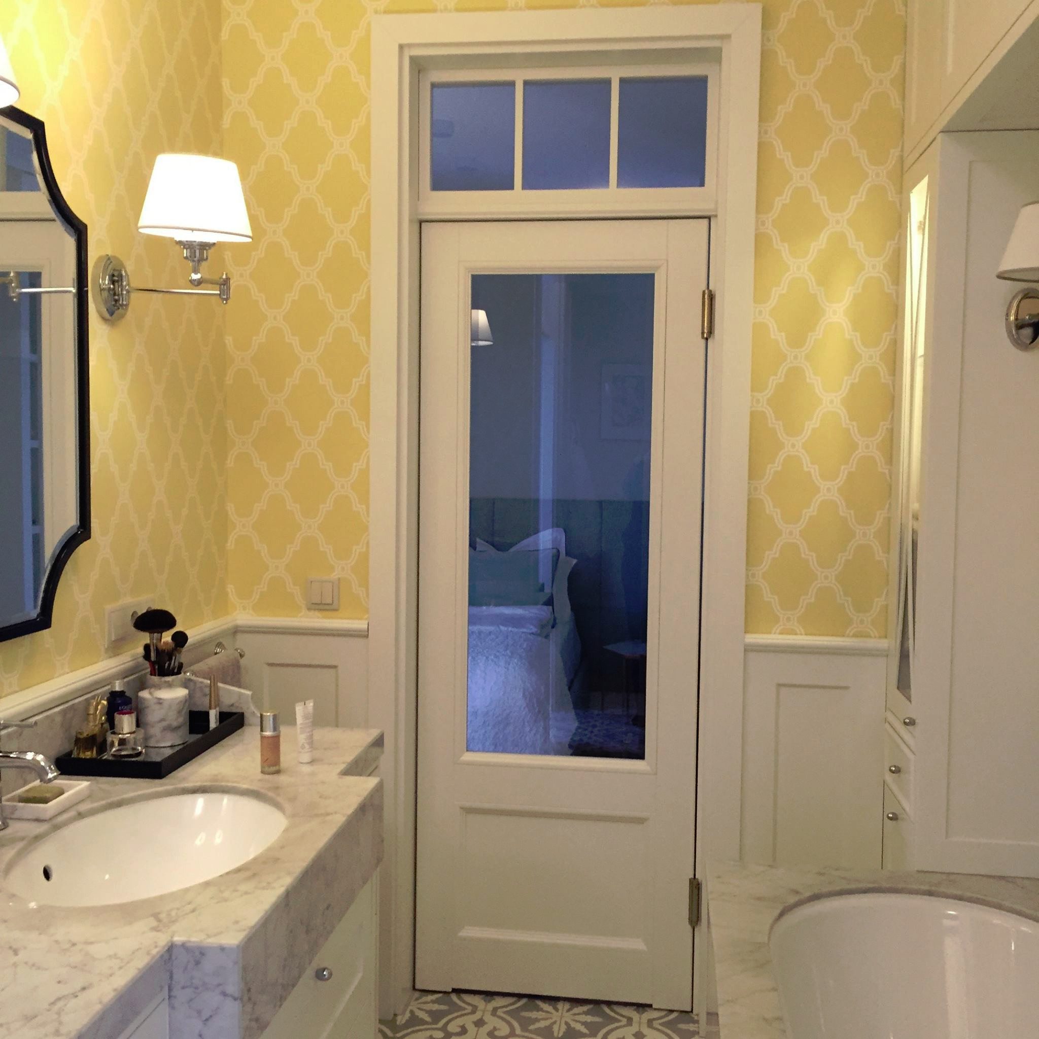 Łazienka z umywalką na marmurowym blacie, żółtą tapetą w białe wzory i przeszklonymi drzwiami do sypialni.