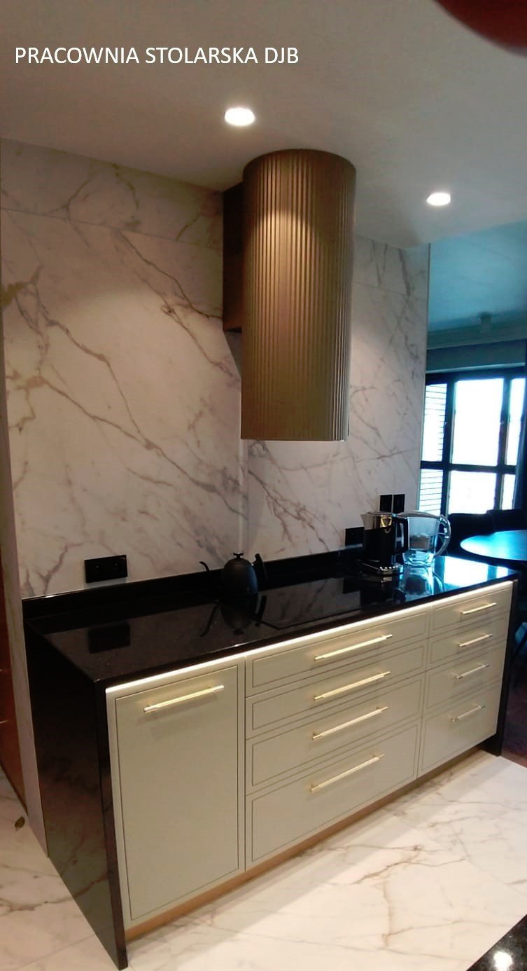 Nowoczesna zabudowa kuchenna z szarymi frontami, złotymi uchwytami i czarnym blatem, uzupełniona o okrągły, metalowy okap w kolorze starego złota na tle marmurowej ściany.