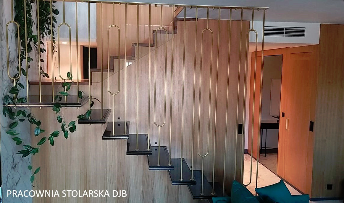 Schody z drewnianymi stopniami i złotą, dekoracyjną balustradą, z elementami roślinnymi, widok na drewnianą ścianę i drzwi.