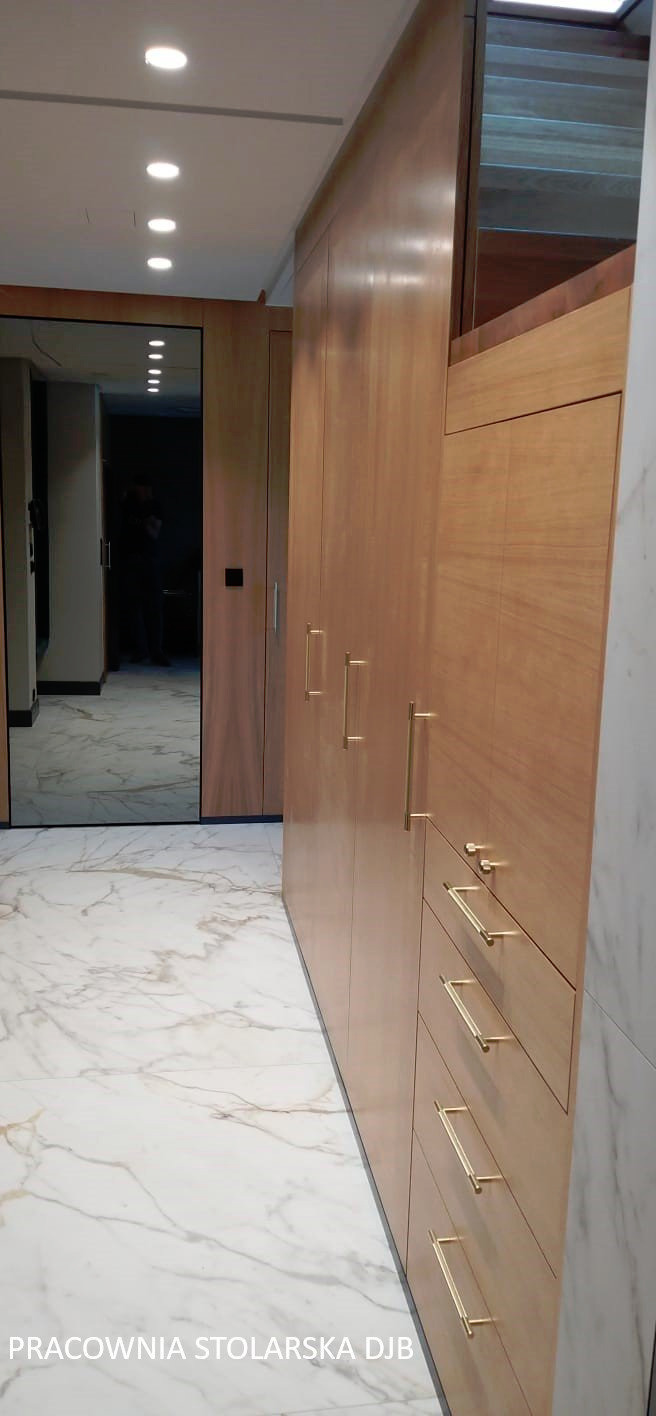 Długa, drewniana szafa z szufladami i złotymi uchwytami, widoczna na tle marmurowej podłogi i lustrzanych drzwi, z logo pracowni stolarskiej w rogu.