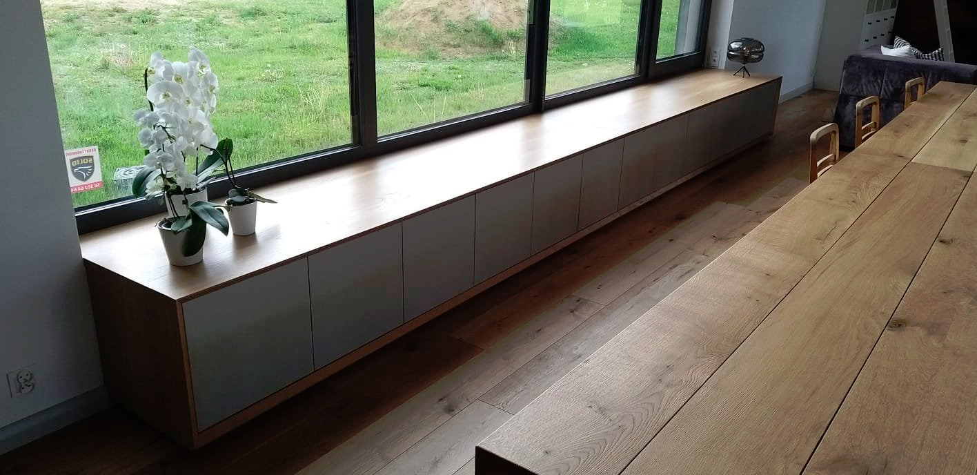 Długa szafka pod oknem z drewnianym blatem i szarymi frontami, na blacie białe storczyki w doniczkach, drewniana podłoga i stół w tle.