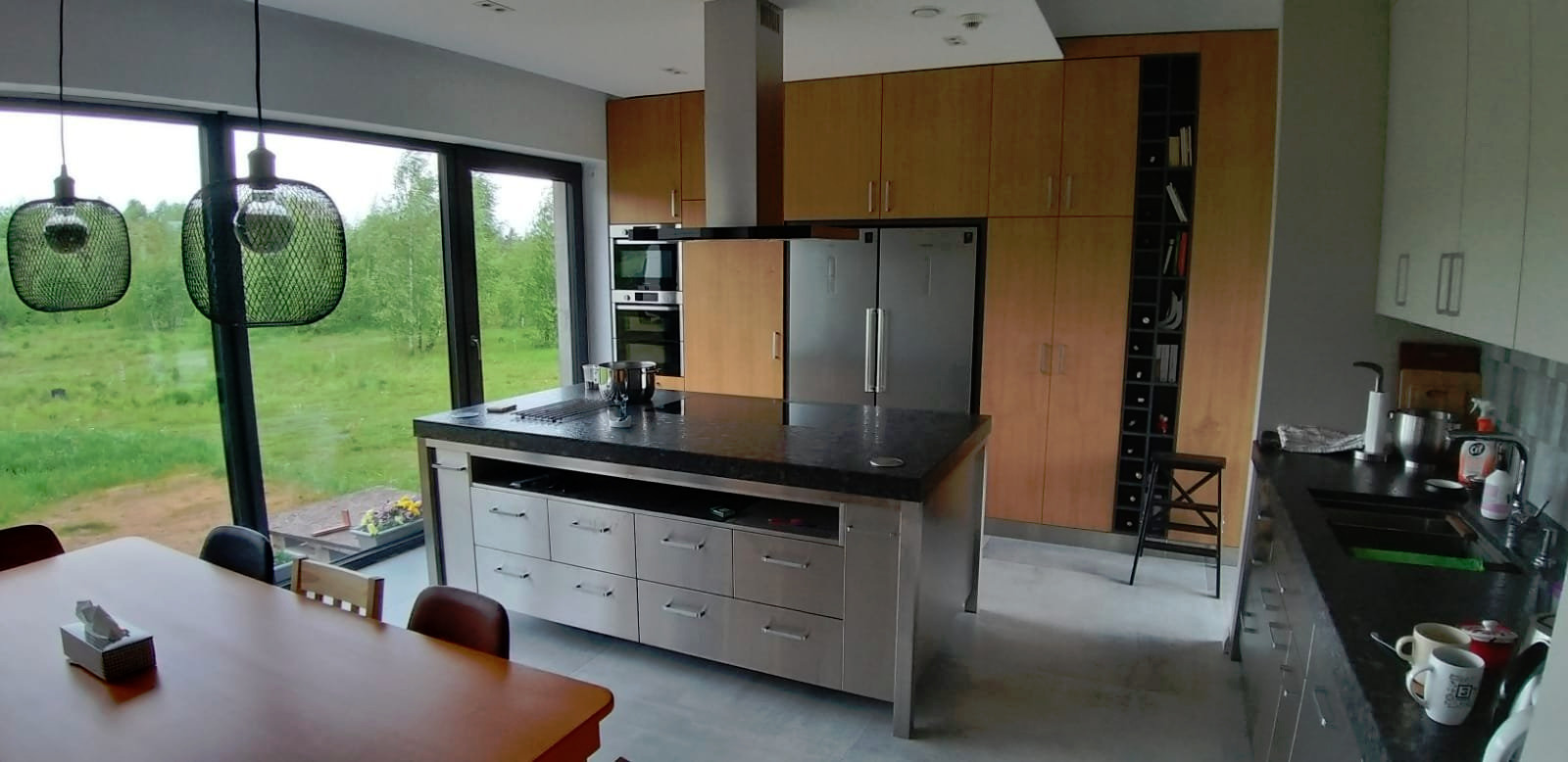 Nowoczesna kuchnia z wyspą na tle dużego okna z widokiem na zieleń, drewniane szafki i stalowe elementy wyposażenia.