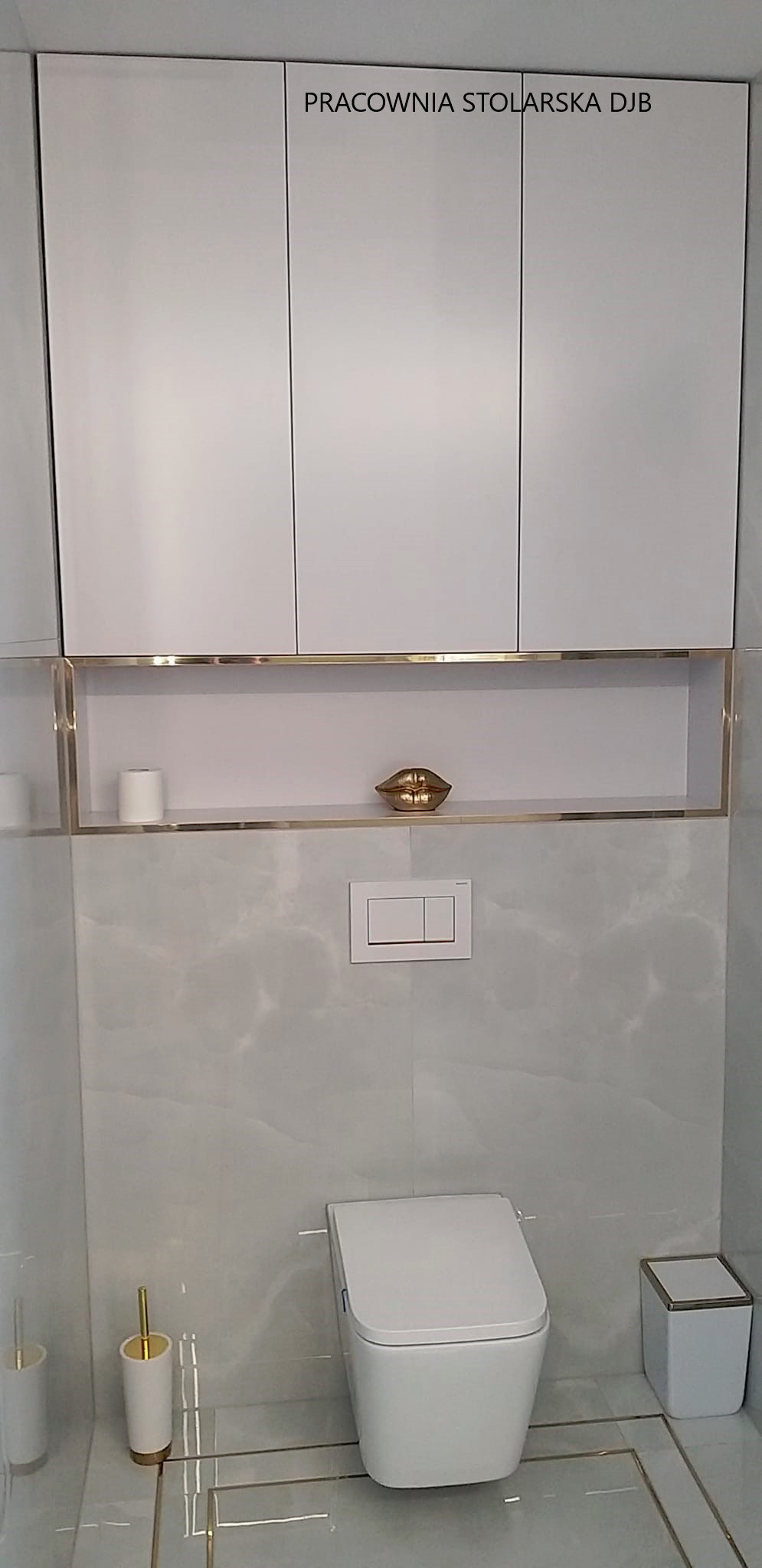 Białe szafki wiszące z otwartą półką z dekoracyjną figurką ust w złotej tonacji nad podwieszaną toaletą w łazience wykończonej marmurowymi płytkami z złotymi akcentami.