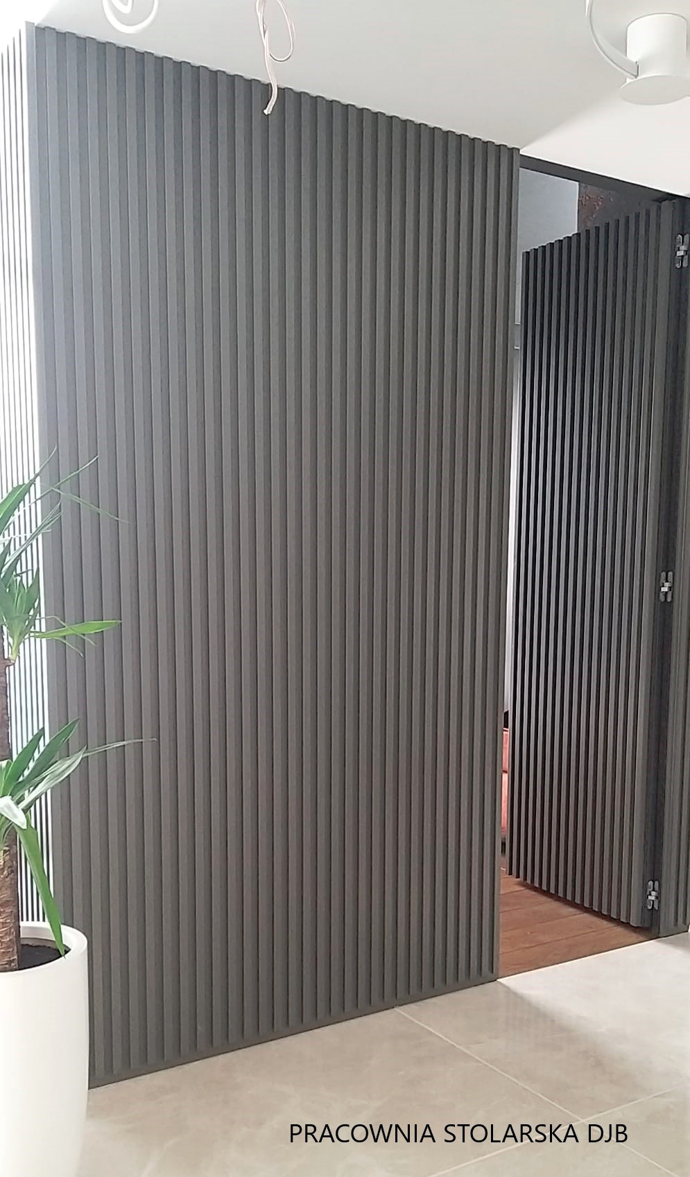 Nowoczesne drzwi harmonijkowe z szarymi lamelami, widoczne z bliska, z fragmentem rośliny doniczkowej w lewym dolnym rogu i fragmentem podłogi z płytek.
