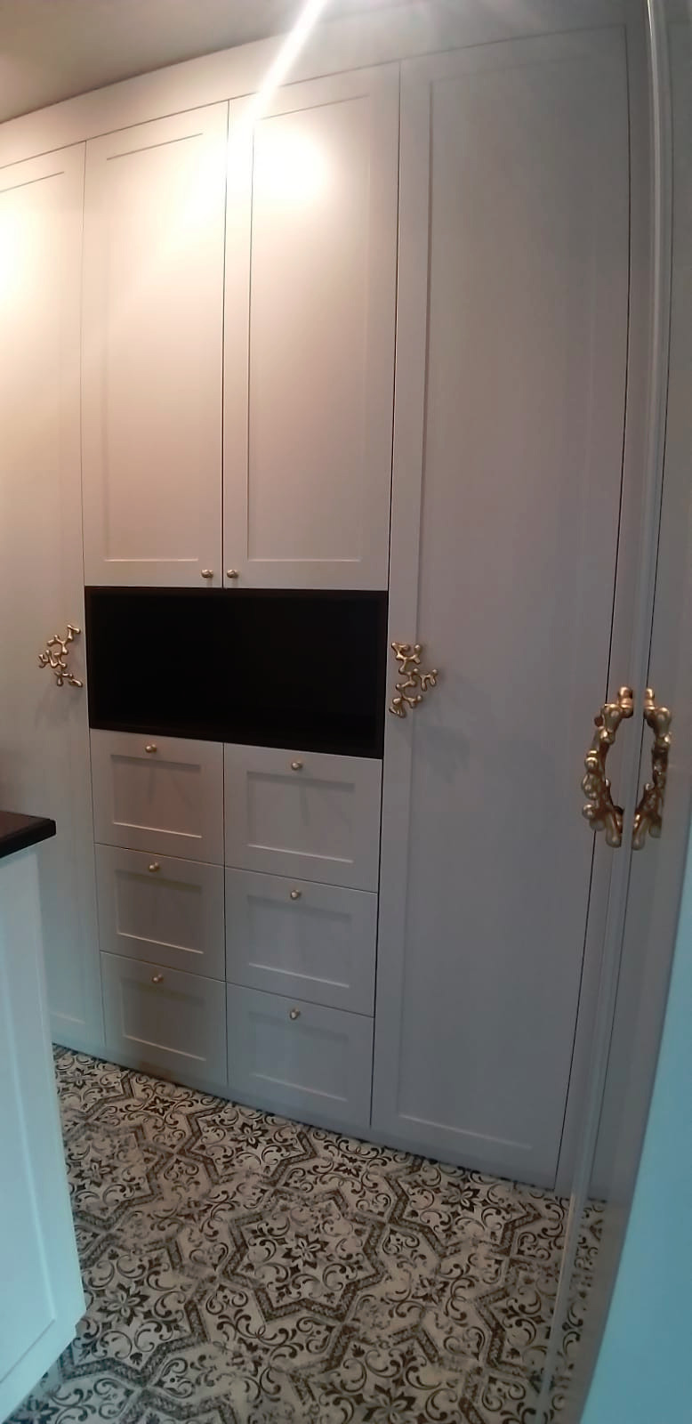 Jasnoszara zabudowa meblowa z szafkami górnymi i dolnymi szufladami, z czarną wnęką i ozdobnymi, złotymi uchwytami na tle wzorzystej podłogi.