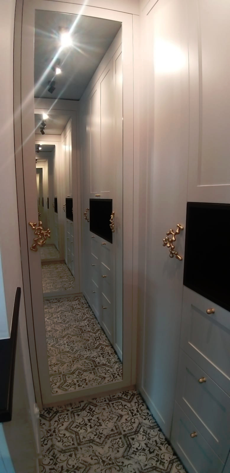 Wąska garderoba z białymi szafami i lustrzanymi drzwiami, nietypowe złote uchwyty, wzorzysta podłoga odbijająca się w lustrze.