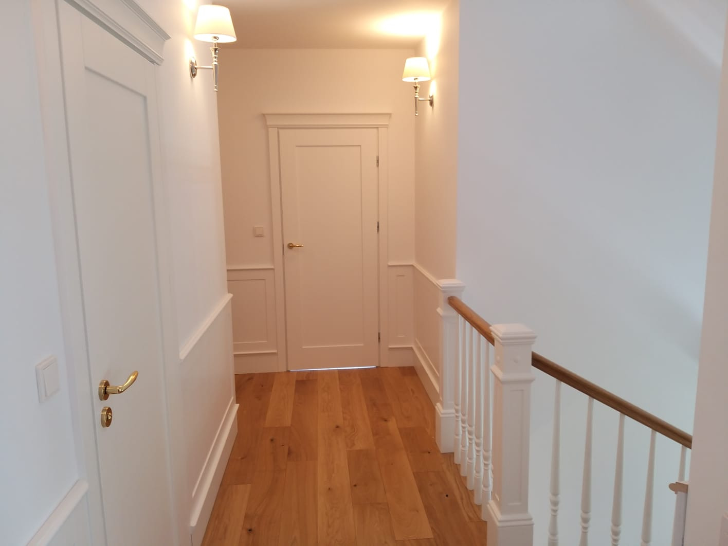 Korytarz z drewnianą podłogą, białymi drzwiami z ozdobnymi listwami, białymi ścianami z lampami ściennymi i białą balustradą schodów z drewnianym poręczą.