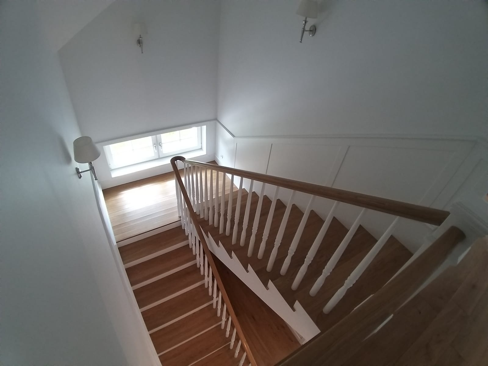 Widok z góry na drewniane schody z białymi balustradami w jasnym pomieszczeniu z oknem i lampami ściennymi.