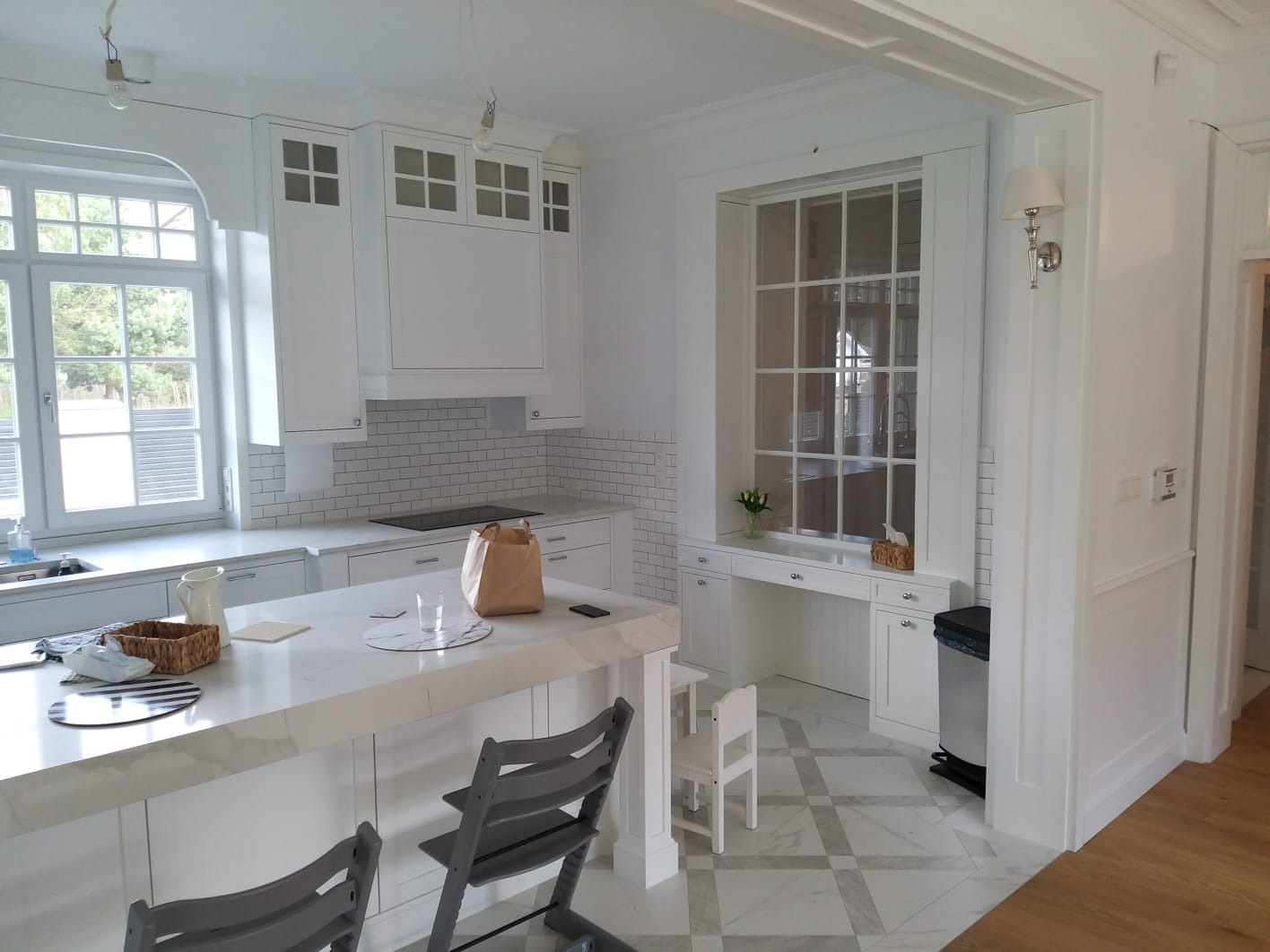 Elegancka, jasna kuchnia w Gdańsku z marmurowym blatem wyspy, białymi szafkami z subtelnymi przeszkleniami i charakterystycznym oknem z zabudową tworzącą miejsce do pracy.