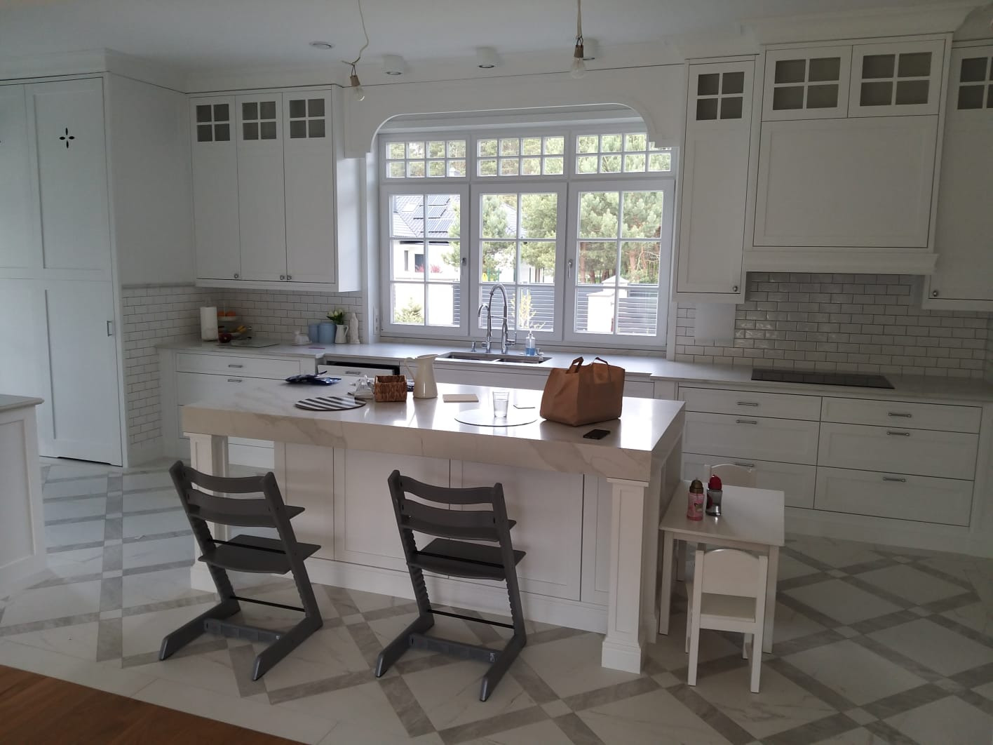 Jasna kuchnia z wyspą, białymi szafkami i blatem z marmuru, okno z widokiem na zieleń, szare krzesła barowe, wzorzysta podłoga.