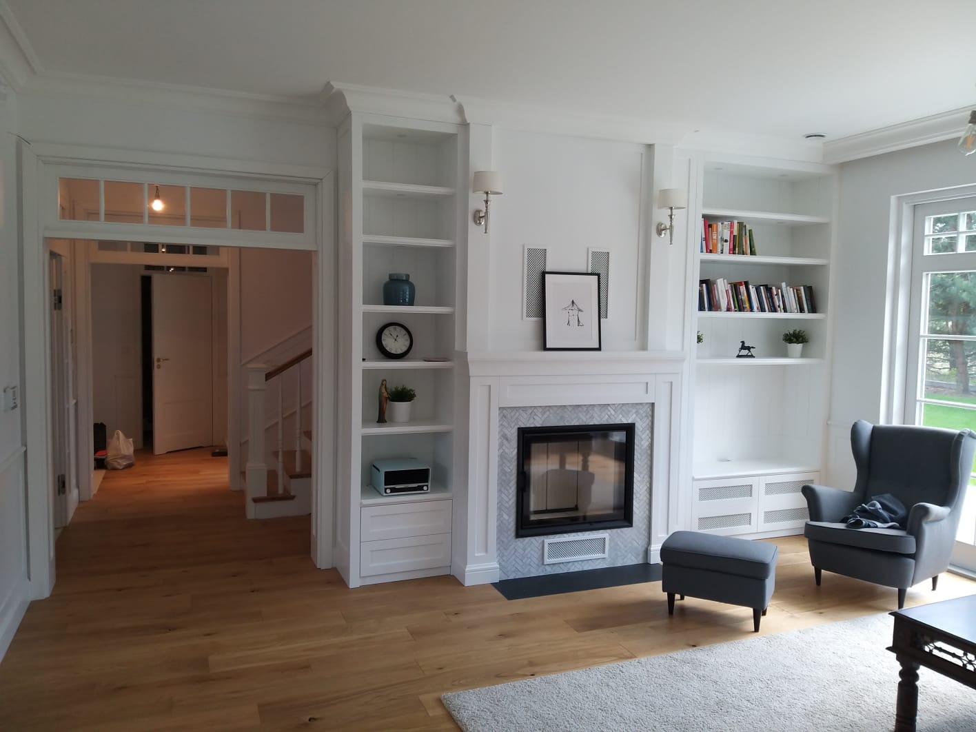 Jasny salon z zabudową biblioteczną po obu stronach kominka z marmurowym wykończeniem, drewniana podłoga i szara sofa z podnóżkiem.