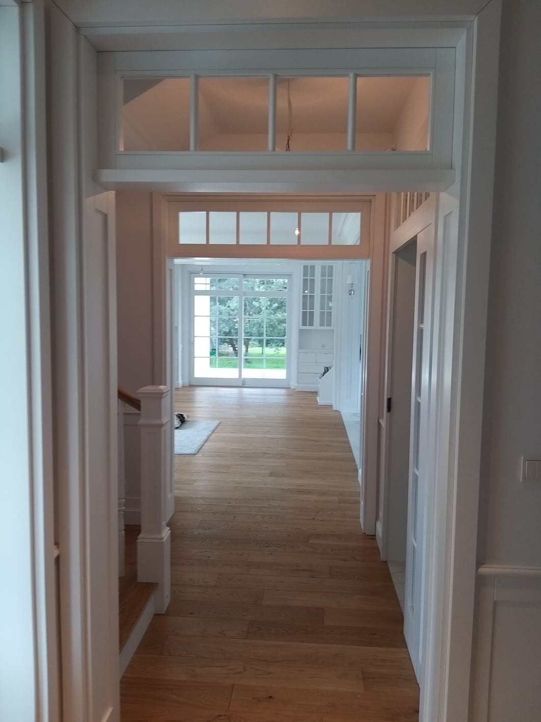 Wnętrze domu z drewnianą podłogą, białymi ścianami z ozdobnymi listwami i białymi drzwiami z przeszkleniami, widok z korytarza na salon