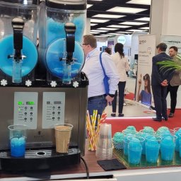 PERSEID SPÓŁKA Z OGRANICZONĄ ODPOWIEDZIALNOŚCIĄ - Automat do mrożonych napojów na stoisku targowym z niebieskimi slushami w kubkach. W tle widoczni ludzie i stoiska.