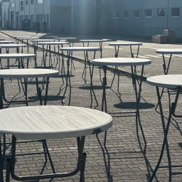 PERSEID SPÓŁKA Z OGRANICZONĄ ODPOWIEDZIALNOŚCIĄ - Rząd białych, okrągłych stolików koktajlowych z metalowymi nogami, ustawionych na zewnątrz na brukowanym placu przed budynkiem. Cienie i ostre światło słoneczne.