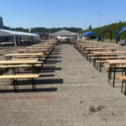 Ustawione rzędy drewnianych stołów i ławek, przygotowane do imprezy plenerowej. W tle białe i niebieskie namioty eventowe.
